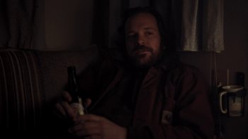 Movie still from “Night Moves” (2013), directed by Kelly Reichardt – A man sitting on a couch holding a bottle of beer; Medium shot, High angle