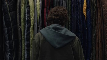 Movie still from “Night Moves” (2013), directed by Kelly Reichardt – A person wearing a jacket and a hood in front of a row of blankets; Medium shot, Overhead angle