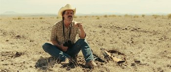 Movie still from “No Country for Old Men” (2007), directed by Joel Coen – A man sitting in the middle of a field holding a camera; Medium shot, Low angle