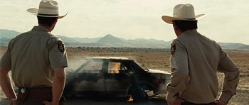 Movie still from “No Country for Old Men” (2007), directed by Joel Coen – A man standing in front of a car on fire; Medium shot, Over the shoulder angle