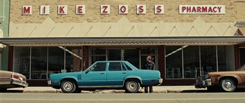 Movie still from “No Country for Old Men” (2007), directed by Joel Coen – A man standing next to a blue car on the side of the street; Wide shot, Over the shoulder angle