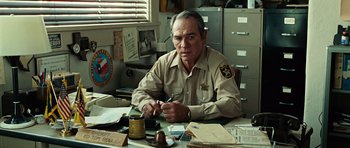 Movie still from “No Country for Old Men” (2007), directed by Joel Coen – A man sitting at a table with a pen in his hand; Medium shot, Over the shoulder angle