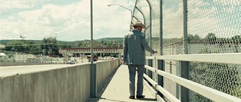 Movie still from “No Country for Old Men” (2007), directed by Joel Coen – A man in a suit and a hat walking on a bridge; Wide shot, Low angle