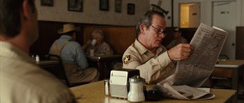 Movie still from “No Country for Old Men” (2007), directed by Joel Coen – An older man sitting at a table reading a newspaper; Medium shot, Over the shoulder angle