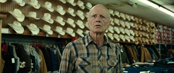 Movie still from “No Country for Old Men” (2007), directed by Joel Coen – An older man wearing a plaid shirt in front of hats; Close Up shot, Over the shoulder angle