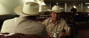 Movie still from “No Country for Old Men” (2007), directed by Joel Coen – Two men in cowboy hats are sitting at a table; Medium shot, Over the shoulder angle