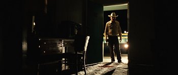 Movie still from “No Country for Old Men” (2007), directed by Joel Coen – A man in a cowboy hat standing in front of an open door; Wide shot, Low angle