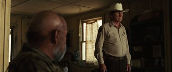 Movie still from “No Country for Old Men” (2007), directed by Joel Coen – An older man wearing a cowboy hat in front of an older man in front of an open window; Medium shot, Over the shoulder angle