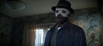 Movie still from “No Sudden Move” (2021), directed by Steven Soderbergh – A man wearing a mask and a hat in a living room; Close Up shot, Over the shoulder angle
