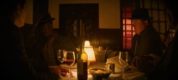 Movie still from “No Sudden Move” (2021), directed by Steven Soderbergh – A man sitting at a table with a bottle of wine and two glasses; Medium shot, Over the shoulder angle