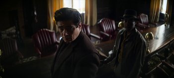Movie still from “No Sudden Move” (2021), directed by Steven Soderbergh – An older man in a brown coat in a room with red chairs; Medium shot, Low angle