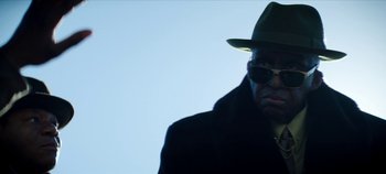 Movie still from “No Sudden Move” (2021), directed by Steven Soderbergh – A man wearing a green hat and a black coat; Close Up shot, Low angle