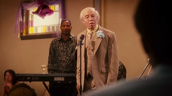 Movie still from “Norbit” (2007), directed by Brian Robbins – An older man wearing a suit standing in front of a microphone; Medium shot, Low angle