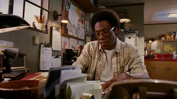 Movie still from “Norbit” (2007), directed by Brian Robbins – A man sitting at a table looking at papers on top of a desk; Medium shot, Low angle