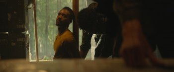 Movie still from “Mother/Android” (2021), directed by Mattson Tomlin – A man sitting in front of a window while another man is standing behind him; Medium shot, Over the shoulder angle