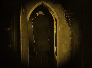 Movie still from “Nosferatu” (1922), directed by F.W. Murnau – A man in a trench coat is standing in an archway; Wide shot, High angle