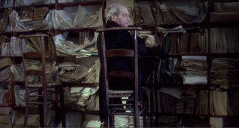 Movie still from “Nosferatu the Vampyre” (1979), directed by Werner Herzog – An older man sitting in a chair in front of a wall of papers; Medium shot, High angle