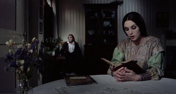 Movie still from “Nosferatu the Vampyre” (1979), directed by Werner Herzog – A woman sitting at a table reading a book while another woman sits in the background; Medium shot, Over the shoulder angle