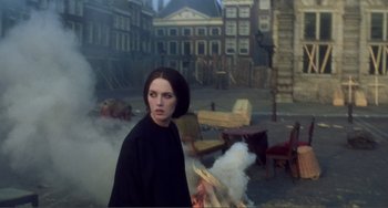 Movie still from “Nosferatu the Vampyre” (1979), directed by Werner Herzog – A woman standing in front of a building with smoke coming out of it; Medium shot, Over the shoulder angle