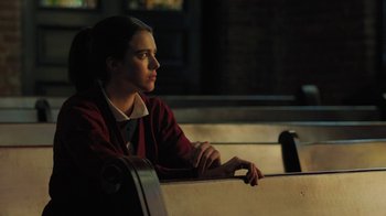Movie still from “Novitiate” (2017), directed by Maggie Betts – A person sitting on a bench in a dark room; Medium shot, Over the shoulder angle