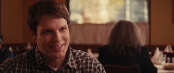 Movie still from “Obvious Child” (2014), directed by Gillian Robespierre – A young man smiling for the camera while sitting at a table; Close Up shot, Over the shoulder angle