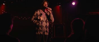 Movie still from “Obvious Child” (2014), directed by Gillian Robespierre – A man holding a microphone while standing on a stage; Medium shot, Over the shoulder angle