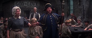 Movie still from “Oliver!” (1968), directed by Carol Reed – A man dressed in a blue suit and hat holding a cane; Medium shot, Over the shoulder angle