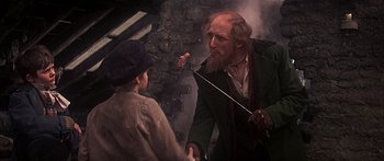 Movie still from “Oliver!” (1968), directed by Carol Reed – An old man with a long white beard is holding a knife and talking to a young boy; Medium shot, Over the shoulder angle