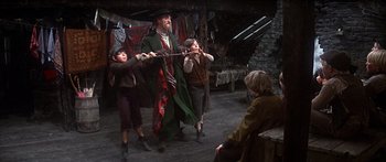 Movie still from “Oliver!” (1968), directed by Carol Reed – A group of people standing on top of a wooden floor; Medium shot, Over the shoulder angle