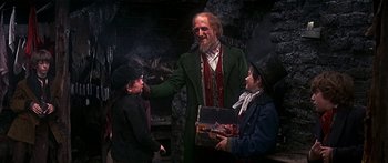 Movie still from “Oliver!” (1968), directed by Carol Reed – A man with a beard is holding a box while two children stand next to him; Medium shot, Over the shoulder angle