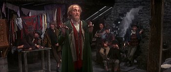 Movie still from “Oliver!” (1968), directed by Carol Reed – An old man with a long beard is holding up his hands in front of a group of people; Medium shot, Over the shoulder angle
