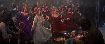 Movie still from “Oliver!” (1968), directed by Carol Reed – A group of women in dresses singing in a crowded room; Wide shot, High angle
