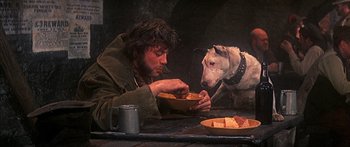 Movie still from “Oliver!” (1968), directed by Carol Reed – A man sitting at a table with a bowl of food and a dog; Medium shot, High angle