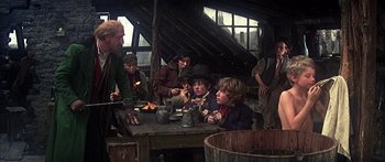 Movie still from “Oliver!” (1968), directed by Carol Reed – A group of people sitting around a table; Medium shot, Over the shoulder angle