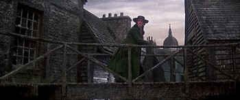 Movie still from “Oliver!” (1968), directed by Carol Reed – A man in a green coat and a hat standing on top of a bridge; Wide shot, Low angle