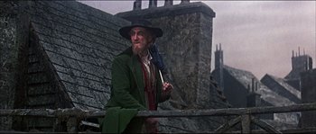 Movie still from “Oliver!” (1968), directed by Carol Reed – A man in a green coat holding an umbrella and sitting on a railing; Medium shot, Low angle
