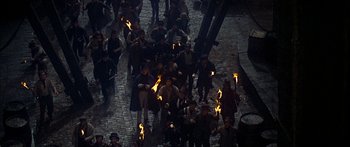 Movie still from “Oliver!” (1968), directed by Carol Reed – A group of men holding torches on a brick road; Wide shot, High angle