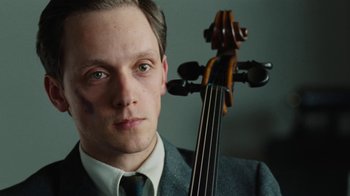 Movie still from “On Chesil Beach” (2017), directed by Dominic Cooke – A man in a suit and tie holding a cello; Close Up shot, Over the shoulder angle