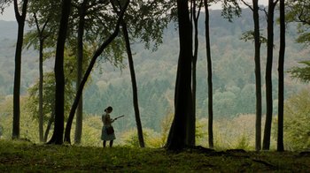 Movie still from “On Chesil Beach” (2017), directed by Dominic Cooke – A woman standing in the middle of a forest holding a guitar; Extreme Wide shot, High angle