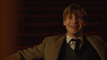 Movie still from “On Chesil Beach” (2017), directed by Dominic Cooke – A man wearing a suit and tie smiling for the camera; Medium shot, Low angle