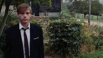 Movie still from “On Chesil Beach” (2017), directed by Dominic Cooke – A man in a suit and tie standing in front of bushes; Medium shot, Over the shoulder angle