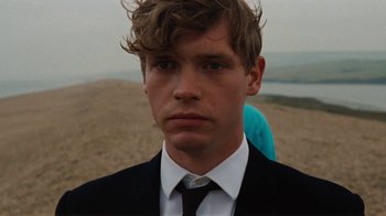 Movie still from “On Chesil Beach” (2017), directed by Dominic Cooke – A man in a suit and tie standing on the beach; Close Up shot, Over the shoulder angle