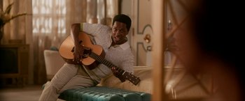 Movie still from “One Night in Miami...” (2020), directed by Regina King – A man sitting on top of a couch holding a guitar; Medium shot, High angle