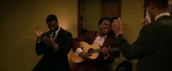 Movie still from “One Night in Miami...” (2020), directed by Regina King – A man in a suit playing a guitar while another man claps in front of him; Medium shot, Over the shoulder angle