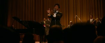Movie still from “One Night in Miami...” (2020), directed by Regina King – A man in a black suit is singing into a microphone in front of a group of people; Medium shot, High angle