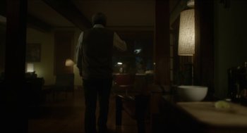 Movie still from “Orphan: First Kill” (2022), directed by William Brent Bell – A man standing in a dark room with a lamp; Wide shot, Over the shoulder angle
