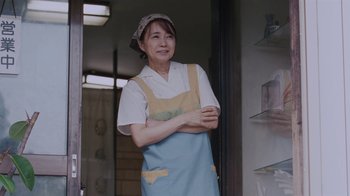 Movie still from “Our Little Sister” (2015), directed by Kore-eda Hirokazu – A woman wearing an apron standing in front of shelves; Medium shot, Low angle
