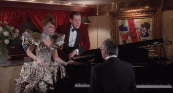 Movie still from “Overboard” (1987), directed by Garry Marshall – A man and a woman are playing piano while a man looks on; Medium shot, Over the shoulder angle