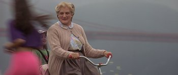 Movie still from “Mrs. Doubtfire” (1993), directed by Chris Columbus – An older woman riding a bike on a cloudy day; Wide shot, Over the shoulder angle
