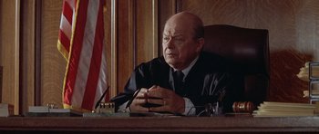 Movie still from “Mrs. Doubtfire” (1993), directed by Chris Columbus – A judge sitting at a table in front of a flag; Close Up shot, High angle